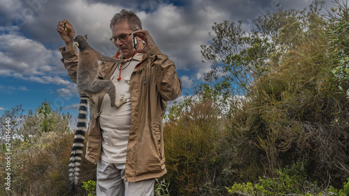 Ring-tailed lemur catta climbs on a man, trying to reach a banana in his hand. The man is smiling, taking pictures on his smartphone. Green vegetation. Blue sky, clouds. Madagascar.  Nosy Soa Park photo