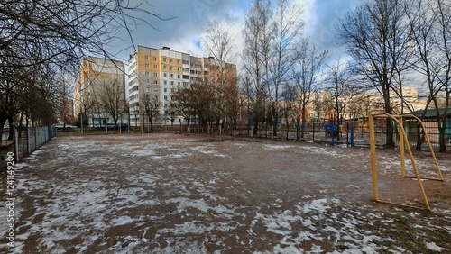 Behind a metal fence surrounded by trees is a stadium with a sandy mini soccer field with goals. Buildings stand nearby. The snow is melting and turning into puddles. The winter sun is breaking photo