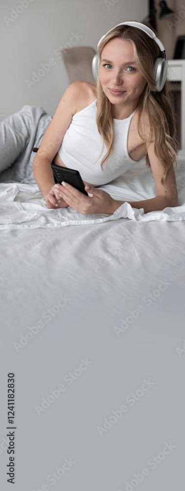 Relaxation technology. Happy student girl wearing wireless headphones using smartphone in bed, enjoying music in bedroom with mobile phone Relaxation technology. Happy student girl wearing wireless headphones using smartphone in bed, enjoying music in bedroom with mobile phone