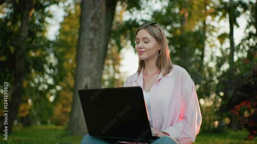 Woman in pink top seated cross-legged on a grassy field, working on her laptop, she looks up thoughtfully with a warm smile, background features trees and greenery, with natural sunlight Woman in pink top seated cross-legged on a grassy field, working on her laptop, she looks up thoughtfully with a warm smile, background features trees and greenery, with natural sunlight