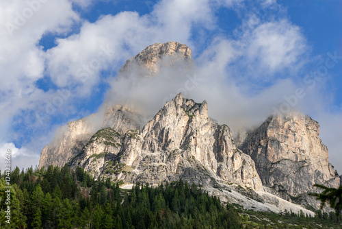 The majestic Sassongher peak in the Dolomites, Italy, rises dramatically above lush forests. The rugged limestone cliffs are partially covered by mist, contrasting against the blue sky photo