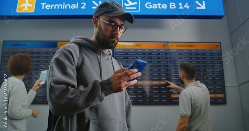 Busy Airport Terminal: Man with Backpack Checks Flight Information Scanning Ticket Using Phone. Traveler Going on Holiday Trip. Digital Arrival and Departure Board for Passengers In the Background.