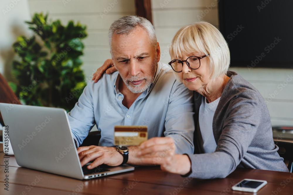 Happy senior married couple using laptop and credit card to make order in online shop while sitting at table at home. Senior couple doing online shopping. Happy senior married couple using laptop and credit card to make order in online shop while sitting at table at home. Senior couple doing online shopping.