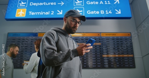 Airport Terminal: Man with Backpack Checks Flight Information Scanning Ticket Using Phone, Looks at Digital Flight Information Display. Tourist Going on Vacation. Diverse People Walking In Background.