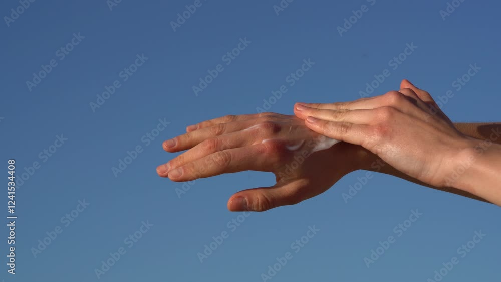 A woman's hand first applies a nourishing cream to her skin and then rubs it in against a blue sky background. The concept of moisturizing and nourishing the skin, natural cosmetics for body care. A woman's hand first applies a nourishing cream to her skin and then rubs it in against a blue sky background. The concept of moisturizing and nourishing the skin, natural cosmetics for body care.