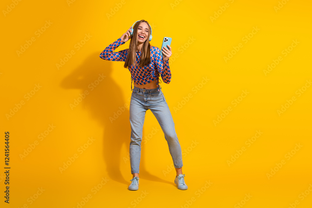 Fototapeta premium Stylish young woman enjoying music on headphones and holding smartphone, standing against vibrant yellow background
