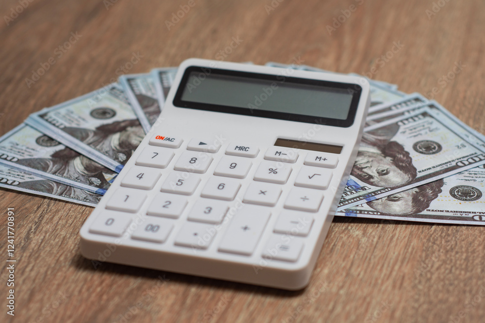 Close-up of a white calculator with money American currency dollars in the background, representing financial calculations. Close-up of a white calculator with money American currency dollars in the background, representing financial calculations.