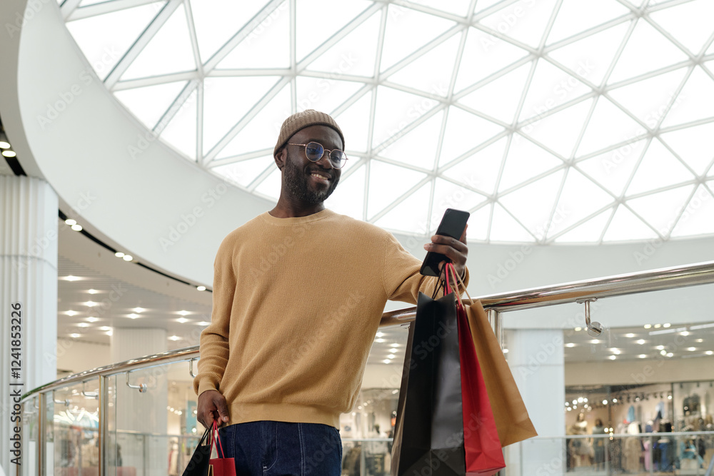 Happy young consumer and blogger with paperbags using smartphone while standing against railings in the mall Happy young consumer and blogger with paperbags using smartphone while standing against railings in the mall