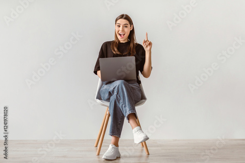 Canvas Print Full length of young Caucasian lady with laptop sitting on chair, pointing upwards, showing IDEA gesture against white wall