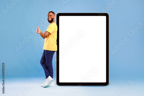 Foto Positive African American guy showing thumb up gesture next to huge digital tablet on blue studio background, empty space for app design on screen
