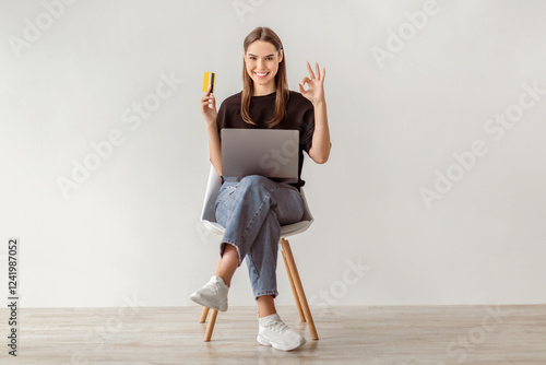 Cybershopping concept. Young lady with laptop and credit card showing okay gesture, sitting on chair against white studio wall, full length. Positive young woman making online purchase on pc photo