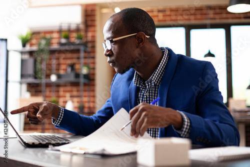 Focused businessman pointing to device screen and reviewing market analysis documents in startup office. Male analyst sitting at desk with laptop and company paperwork, preparing project presentation.