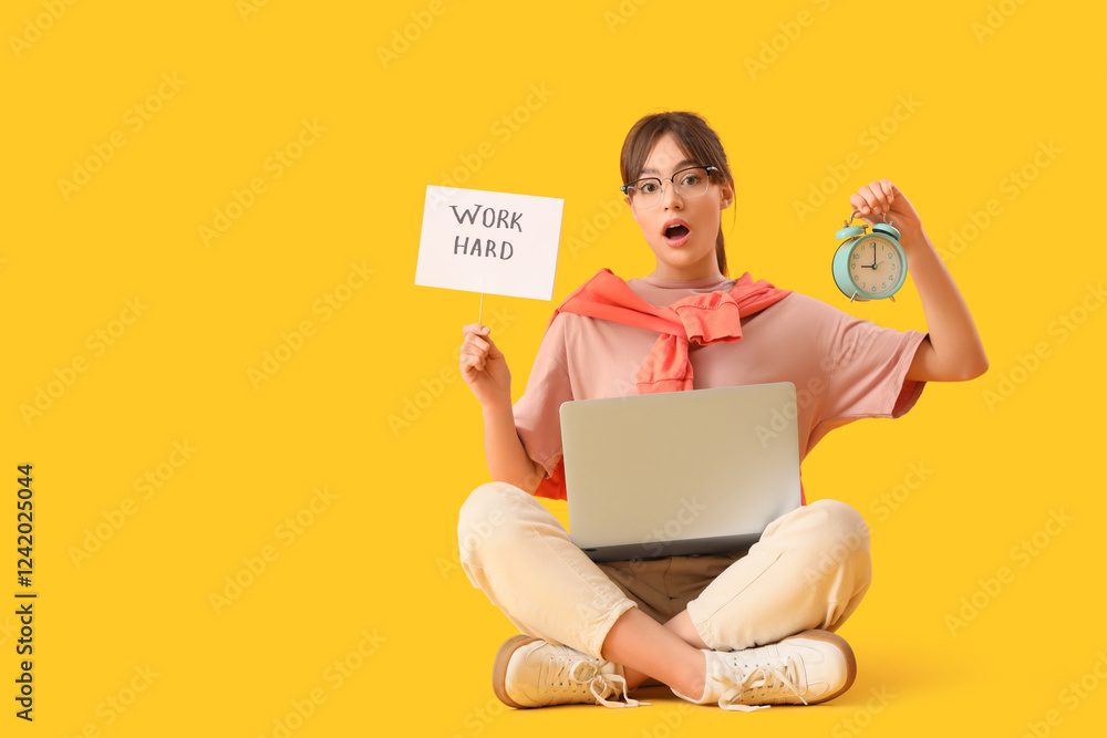 Shocked teenage girl holding paper with text WORK HARD and alarm clock on yellow background Shocked teenage girl holding paper with text WORK HARD and alarm clock on yellow background