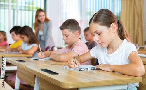 Portrait of concentrated tween girl studying in classroom, listening to schoolteacher and writing in notebook