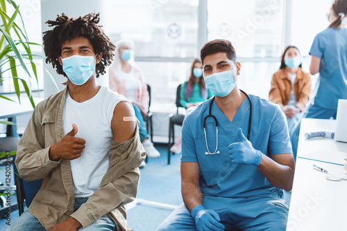 Safety, Mass flu Vaccination. Vaccinated African American Guy Patient And Medical Worker Gesturing Thumbs-Up After Successful Coronavirus Immunization Wearing Face Masks In Health Centre Interior photo