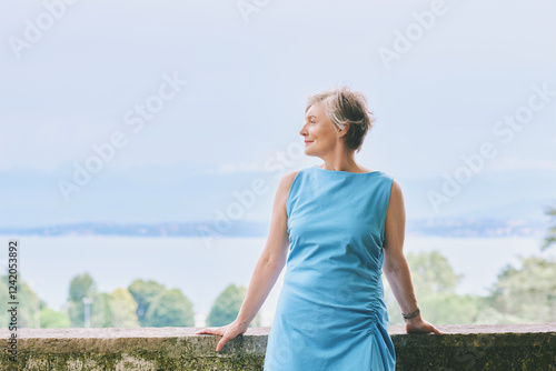 Outdoor portrait of beautiful mature woman of 50 - 55 years old, wearing blue dress, enjoying nice day next to water