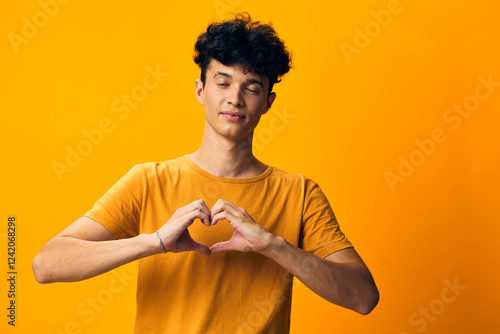 young man making heart shape with hands, smiling, against bright yellow background, expressing love and positivity in a cheerful manner, ideal for romantic themes or friendship concepts photo