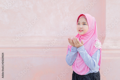 Young Asian Muslim woman in pink hijab and blue striped sleeves praying against soft pink background. Modern modest fashion, religious devotion concept.