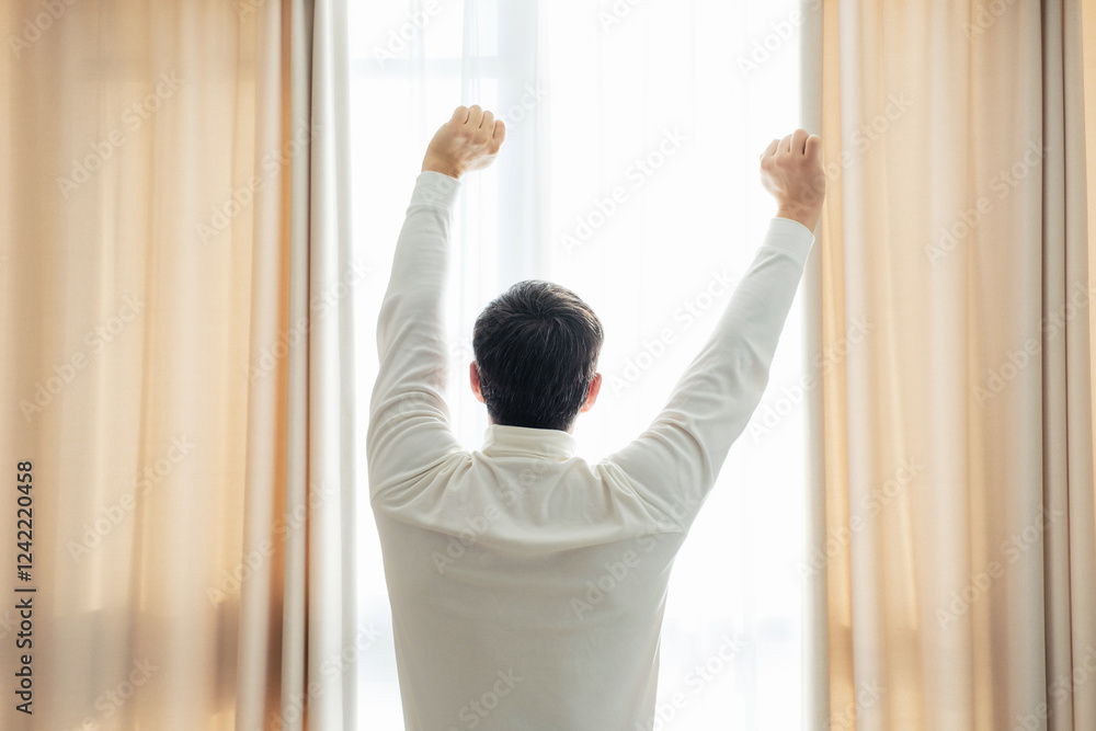Man standing by window raising hands up and stretching, concept of early awakening.