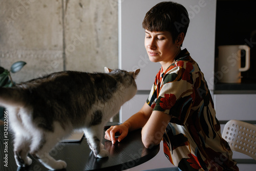 Woman looking at cat walking on table with laptop at home kitchen photo