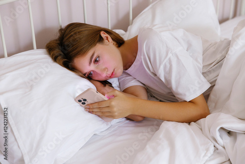 Teenage girl using smartphone lying under blanket on bed at home photo