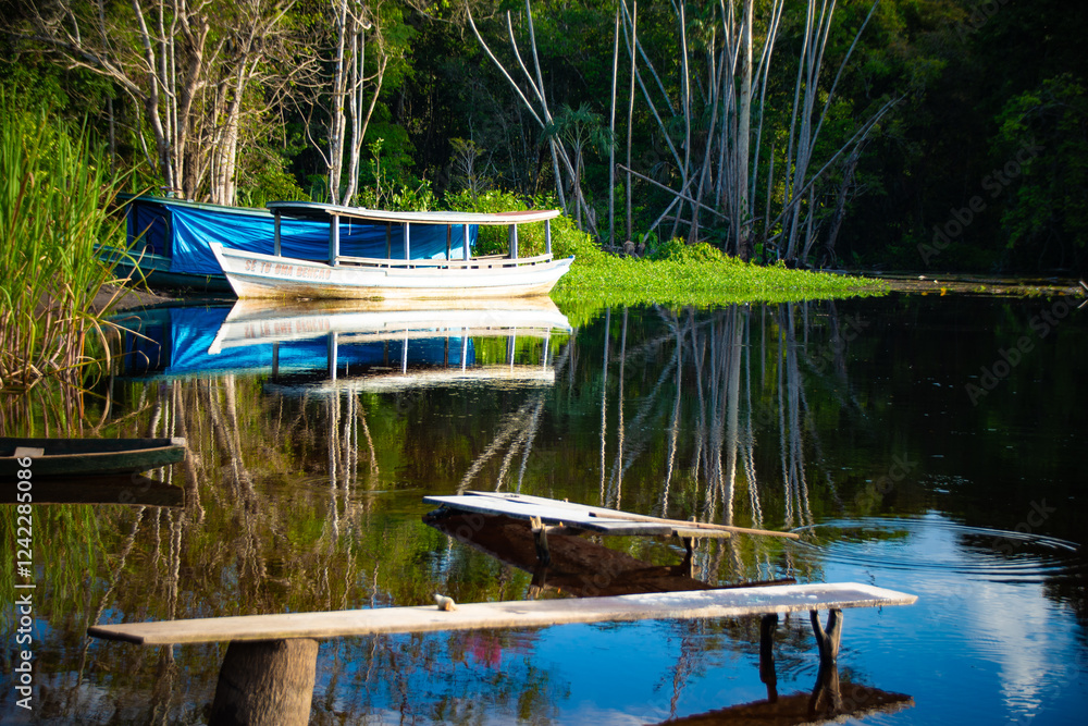 Small wooden boat anchored on a calm river with a larger white boat covered with a blue tarp in the background, surrounded by dense vegetation reflected in the water.
