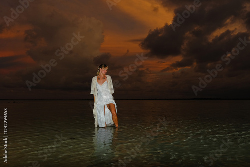 Woman in white dress stands in shallow water at sunset photo