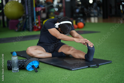 Asian man wearing white headphones stretching while in gym.