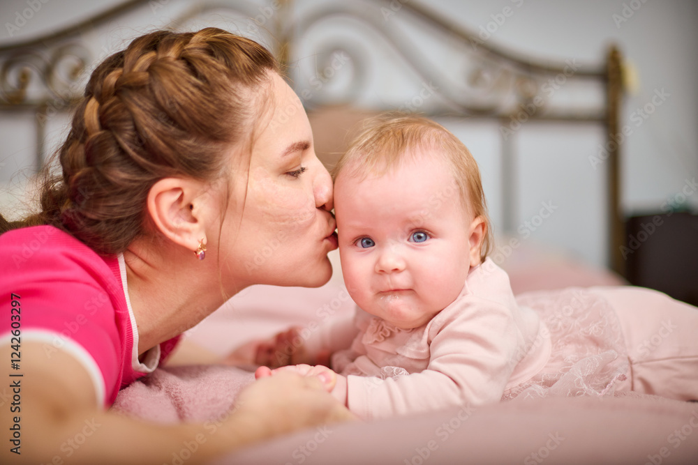 Young mother with braided hair kisses light-haired baby girl on pink bedspread, expressing warmth and love. Soft lighting in cozy bedroom setting enhances tenderness Young mother with braided hair kisses light-haired baby girl on pink bedspread, expressing warmth and love. Soft lighting in cozy bedroom setting enhances tenderness
