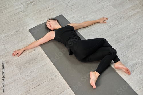 Foto Young adult woman with light skin in black activewear practices supine twist yoga pose on gray mat