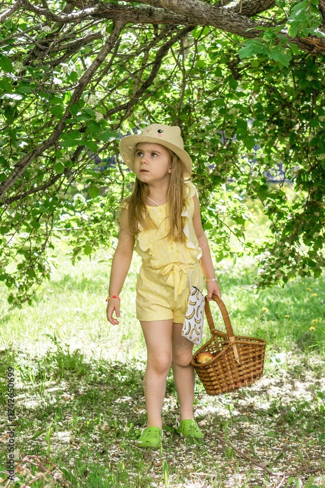 Obraz premium Serious little girl in a yellow dress and straw hat carrying a heavy basket lost in the woods.