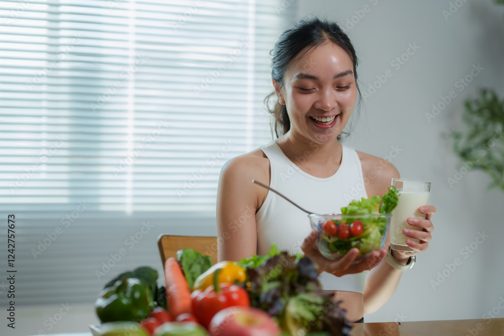 Young asian sportswoman holding a bowl of fresh vegetable salad and a glass of milk while smiling and sitting at the table in the dining room, promoting a healthy lifestyle and clean food concept Young asian sportswoman holding a bowl of fresh vegetable salad and a glass of milk while smiling and sitting at the table in the dining room, promoting a healthy lifestyle and clean food concept