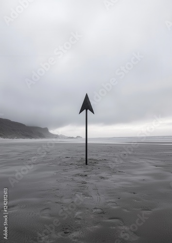 Black and white photograph of a beach with a triangular signpost in the center. the signpost is made of metal and has a pointed top. the beach is covered in sand and there are footprints in the sand. photo