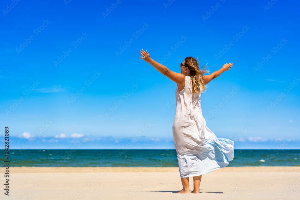 Beautiful middle aged woman in dress fluttering on wind standing with arms raised up on white sandy beach on sunny summer day. Back view. Freedom and energy of life Beautiful middle aged woman in dress fluttering on wind standing with arms raised up on white sandy beach on sunny summer day. Back view. Freedom and energy of life