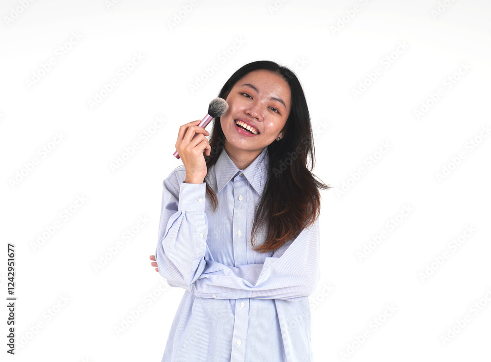 Smiling asian woman with one folded arm wearing blue shirt applying blush make up with pink brush isolated on horizontal white background.