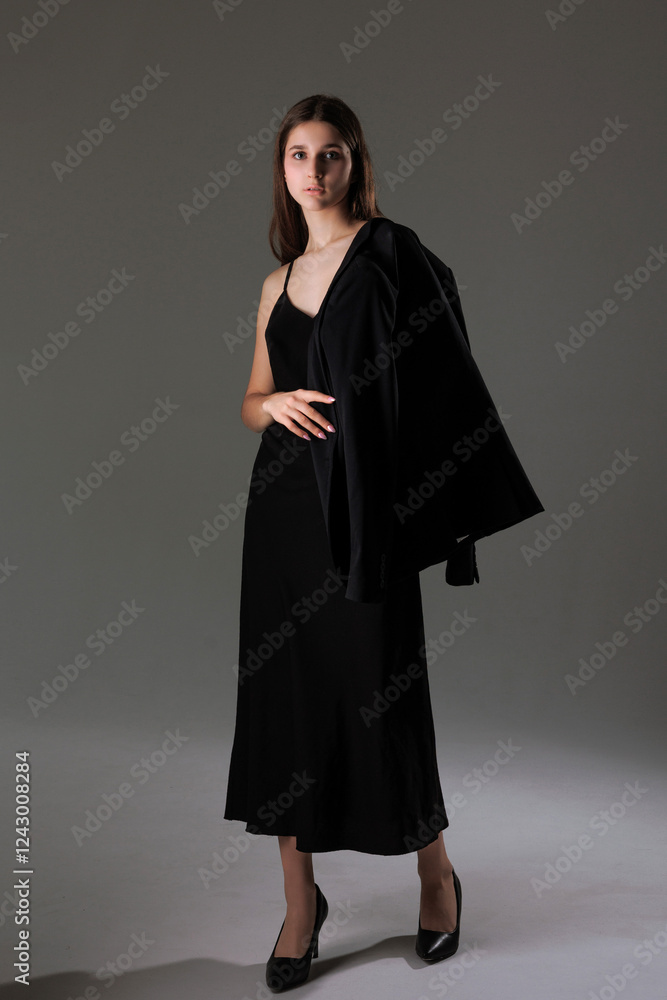 Fototapeta premium Elegant young woman poses in a sleek black dress and jacket in a minimalist studio setting