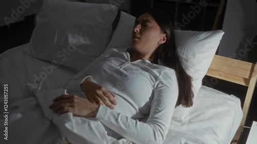 Woman experiencing a nightmare in a dimly lit bedroom at night, wearing a white shirt and covered with a blanket, showing discomfort and unease indoors.