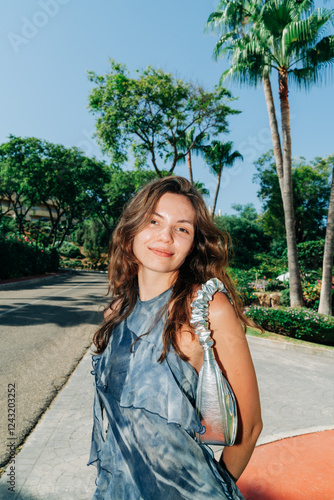 A smiling woman in a blue flowing dress and metallic bag poses on a sunny day surrounded by lush greenery and palm trees
