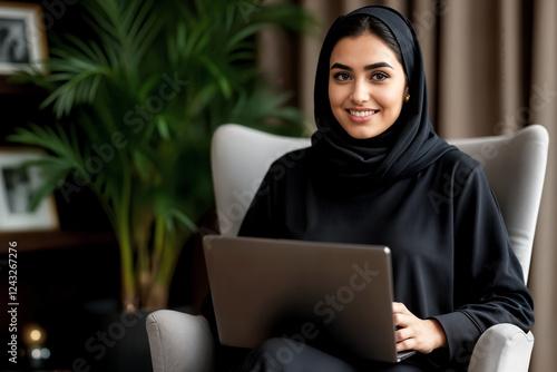 A confident young woman wearing a black hijab sits in a modern, cozy office space, smiling while working on her laptop. She is seated in a comfortable chair, surrounded by elegant decor and greenery photo