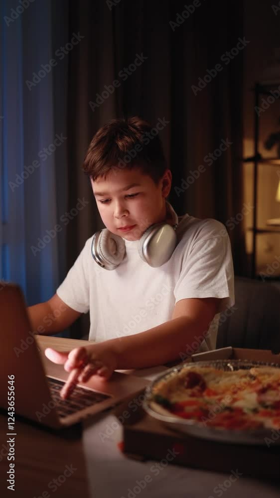 Vertical video of little multiethnic boy at night working on homework typing browsing having distance education on laptop computer and eating pizza fast food at home Vertical video of little multiethnic boy at night working on homework typing browsing having distance education on laptop computer and eating pizza fast food at home