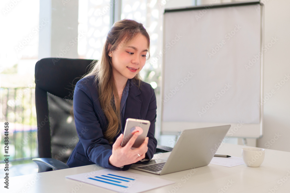 Young Asian businesswoman holding a smartphone while working on a laptop in a modern office with a presentation board Young Asian businesswoman holding a smartphone while working on a laptop in a modern office with a presentation board