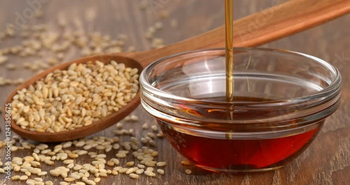 Roasted sesame oil is poured into a glass bowl on a wooden spoon with roasted sesame seeds in the background.