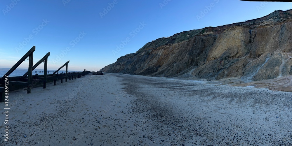 Obraz premium Panoramic landscape view of beautiful sandy beach with blue skies and wooden defence barrier structure pools of sea water reflect sky and chalk cliffs in background at sundown in Winter in Norfolk uk