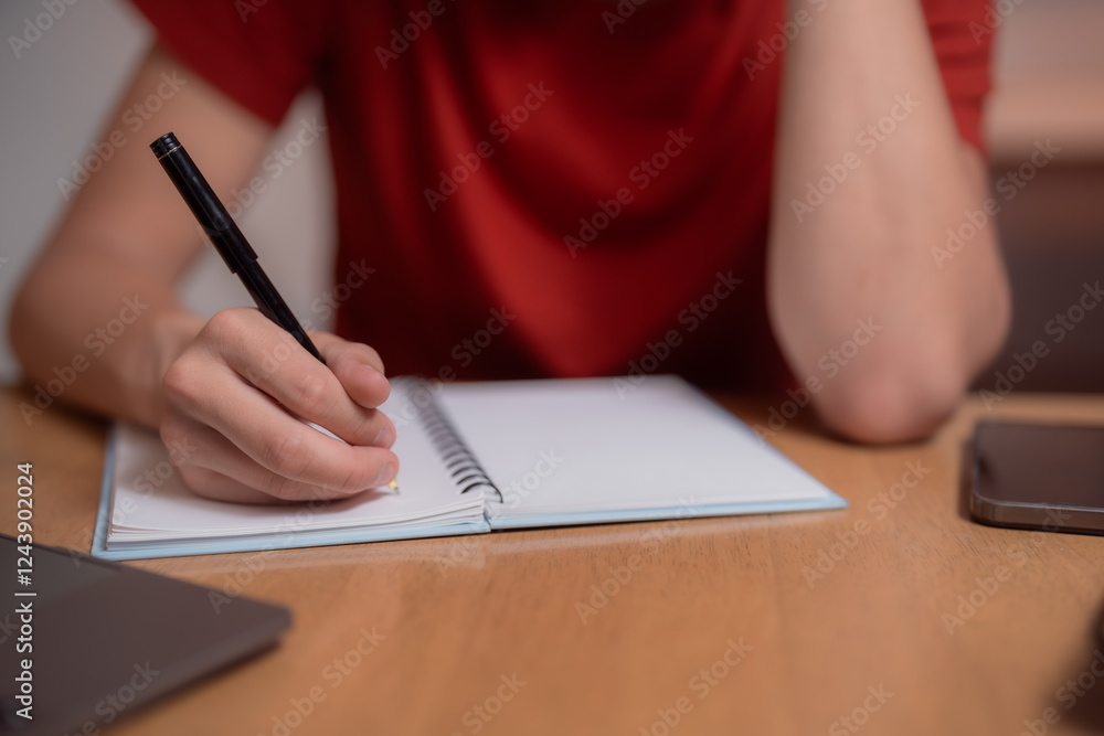 Fototapeta premium Close-up of a student's hand writing in a notebook with a pen, highlighting the act of note-taking and studying, with a smartphone and laptop nearby on the wooden desk