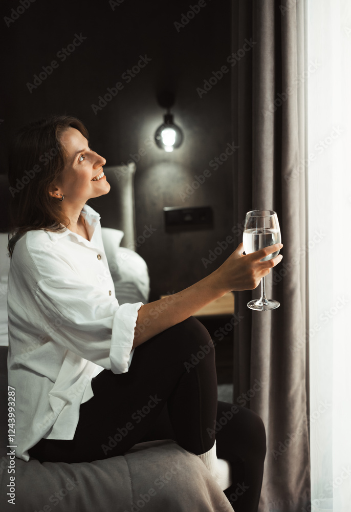 Woman sits on the edge of cozy comfortable hotel bed, looking out the window dreaming with glass of water. Person reflecting on travel experiences and plans, surrounded by her luggage. Woman sits on the edge of cozy comfortable hotel bed, looking out the window dreaming with glass of water. Person reflecting on travel experiences and plans, surrounded by her luggage.