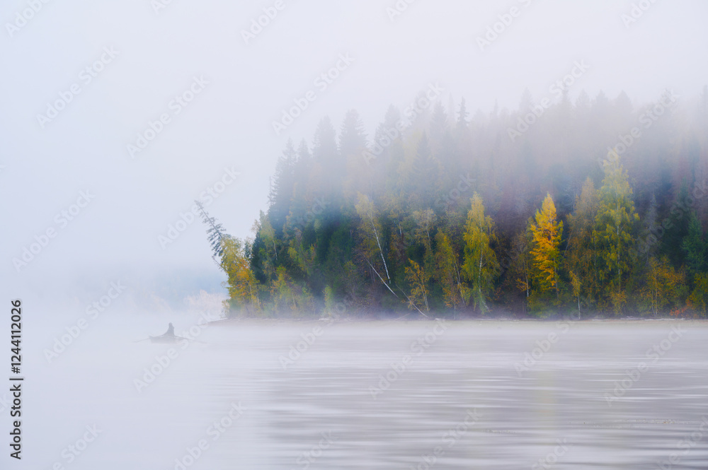 Fototapeta premium Autumn's soft landscape, with morning fog hovering over the river. A fisherman in his boat, surrounded by a colorful forest.