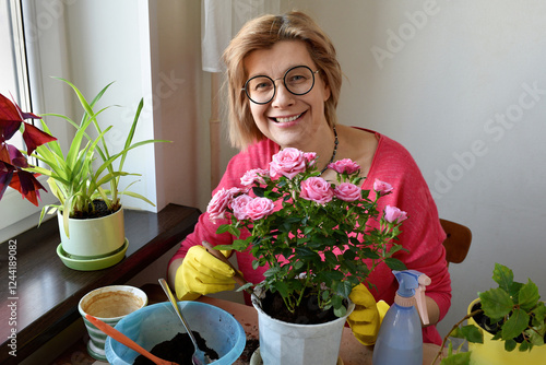 Happy smiling middle aged woman replanting houseplant, blooming rose. Slow life, hobby, happiness.