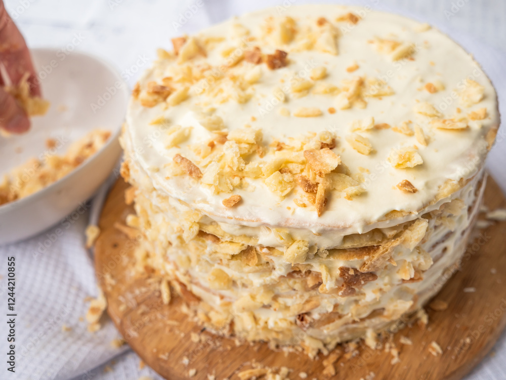 A hand sprinkling decorating crumbs over a pile of thin round layers spread with vanilla custard, a bowl of dough crumbs on a waffle towel in blurry background. Shortcrust puff pastry Napoleon cake.