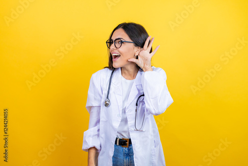 Young brunette doctor woman wearing stethoscope standing over isolated yellow background surprised with hand over ear listening an hearing to rumor or gossip