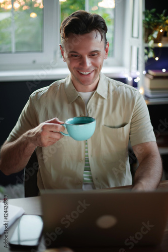 A young man smiles while holding a cup of coffee in one hand and working on his laptop. The photo is set in a cozy workspace with natural light and plants, creating a relaxed, productive vibe. photo